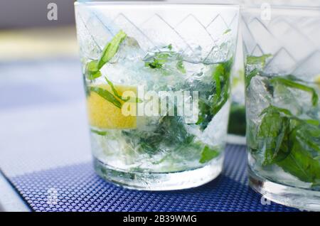 Komplettes Glas frisches, kühles, transparentes Wasser mit Eis, Zitrone und, die Blätter auf blauem Tisch liegen lassen Stockfoto