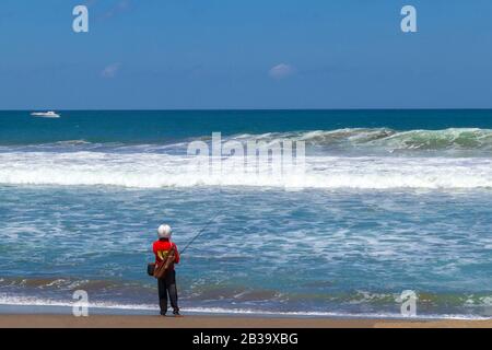 Fischer im Helm sieht aus wie Astronaut steht und angeln Der Strand neben dem Meer Stockfoto