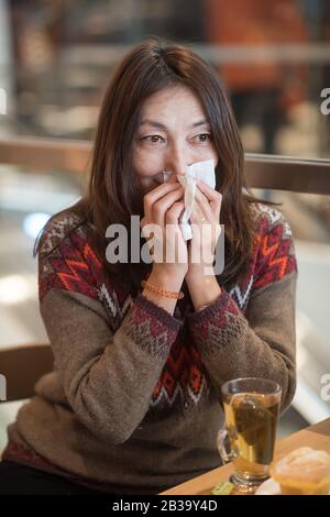 Nette asiatische junge Frau, die nach dem Einkaufen in einem Café die Nase wischt und eine Tasse Tee hat Stockfoto