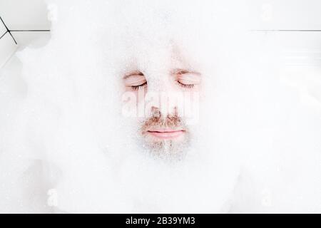 Männliches Gesicht in einem üppigen weißen Schaum, nur Augen und Mund sind sichtbar, Nahaufnahme. Der Mann entspannt sich, während er in einer Badewanne liegt, mit seifigem Wasser. Stockfoto