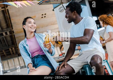 Selektiver Fokus des fröhlichen asiatischen Mädchens, das in der Nähe des afroamerikanischen Mannes lächelt, während man Flaschen mit Bier klinkt Stockfoto