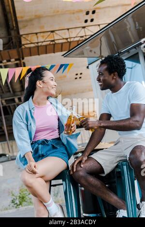 Fröhliches asiatisches Mädchen lächelt in der Nähe des afroamerikanischen Mannes und klinkende Flaschen Bier in der Nähe des Lebensmittel-Trucks Stockfoto