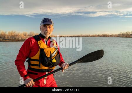 Umweltportrait eines männlichen Paddlers in einem Trockenanzug und einer Rettungsweste, die ein Stehpaddel an einem Seeufer hält Stockfoto