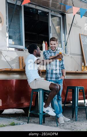 Fröhliche multikulturelle Männer klinken Flaschen mit Bier in der Nähe von Lebensmittel-Truck Stockfoto