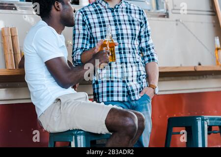 3/4-Blick auf multikulturelle Männer, die Flaschen mit Bier in der Nähe von Lebensmittel-Trucks klinken Stockfoto