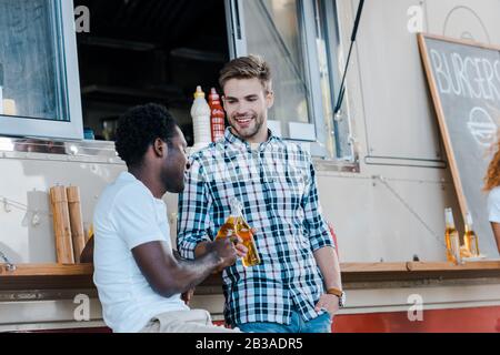 Fröhliche multikulturelle Männer klinken Flaschen mit Bier in der Nähe von Speisewagen Stockfoto