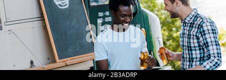 Panoramaaufnahme von glücklichen multikulturellen Männern, die Flaschen Bier in der Nähe von Speisewagen draußen halten Stockfoto