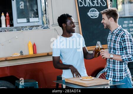 Lächelnde multikulturelle Freunde, die Flaschen Bier in der Nähe von pommes frites und Essenswagen toben