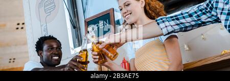 Panoramaaufnahme von fröhlichen multikulturellen Männern, die mit Bierflaschen mit attraktiver Rotkopffrau in der Nähe von Lebensmittel-Lastwagen klinken Stockfoto