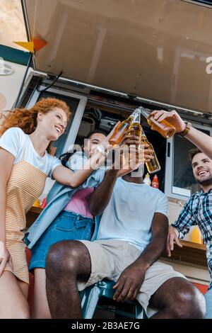 Niedriger Blickwinkel auf glückliche multikulturelle Freunde, die Flaschen mit Bier in der Nähe von Lebensmittel-Trucks klinken Stockfoto