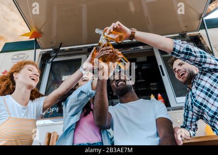 Niedriger Blickwinkel auf fröhliche multikulturelle Freunde, die Flaschen mit Bier in der Nähe von Lebensmittel-Trucks klinken Stockfoto