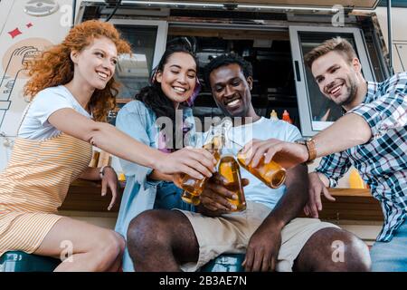 Fröhliche multikulturelle Freunde klinken Flaschen mit Bier in der Nähe von Speisewagen Stockfoto