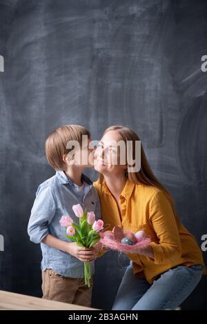 Süßer Sohn, der Mütter küsst, überprüft und ihr Tulpen gibt, während er sie mit Ostern feiert Stockfoto