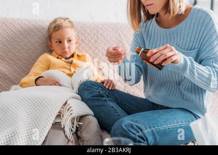 3/4-Ansicht der Mutter, die Hustensirup im Löffel gießt, während sie in der Nähe eines kranken Sohnes sitzt Stockfoto