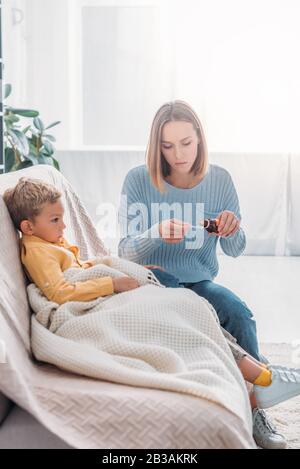 Aufmerksame Mutter schüttet Hustensirup im Löffel, während sie in der Nähe eines kranken Sohnes sitzt Stockfoto