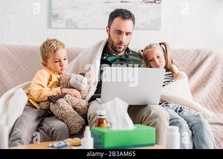 Erkrankte Kinder, die mit einem Laptop auf dem Sofa in der Nähe eines kranken Vaters sitzen Stockfoto
