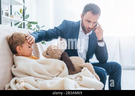 Besorgter Vater, der die Stirn eines kranken Sohnes berührt, während er auf dem Smartphone spricht Stockfoto