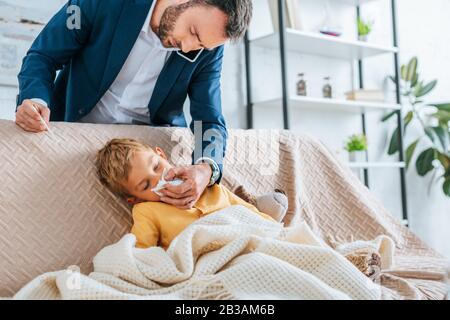 Aufmerksamer Vater wischt die Nase des kranken Sohnes, während er Thermometer hält und auf dem Smartphone spricht Stockfoto