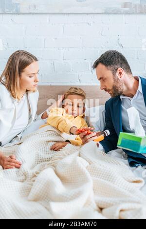 Besorgter Vater, der Hustensirup hält und Mutter den Kopf des erkrankten Sohnes berührt, der im Bett liegt Stockfoto