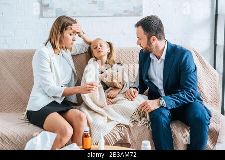 Besorgte Mutter, die die Stirn berührt und beim Sitzen in der Nähe erkrankter Tochter und Ehemann Thermometer betrachtet Stockfoto