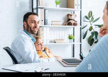 Lächelnder Kinderrist umarmend Junge beim Blick auf die Mutter, die am Schreibtisch sitzt Stockfoto