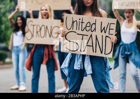 3/4-Ansicht des Mädchens, das Plakat mit der Aufschrift united hält, wir stehen in der Nähe von Feministinnen auf der Straße Stockfoto