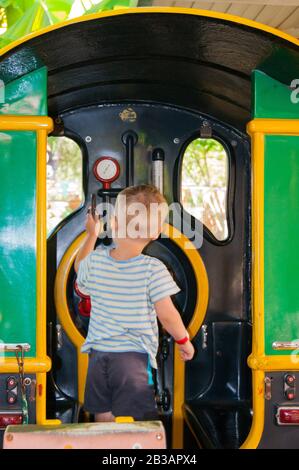 Ein kleiner Junge von drei Jahren reitet in einem Kinderzug im Vergnügungspark Riviera, Sotschi mit dem Rücken zum Zuschauer. Ruhe, Tropen, Ruhe, Aktivierung. Vertikales Foto Stockfoto