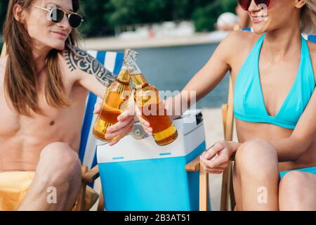 Junger Mann und Frau klinken Flaschen Bier, während sie in Chaiselounges in der Nähe eines tragbaren Kühlschranks sitzen Stockfoto