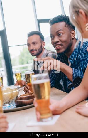 Selektiver Fokus des fröhlichen afroamerikanischen Mannes, der mit dem Finger zeigt, während er mit multikulturellen Freunden in der Kneipe sitzt Stockfoto