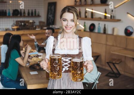 Attraktive Kellnerin in traditioneller deutscher Tracht, die Tassen helles Bier hält und mit der Kamera lächelt Stockfoto