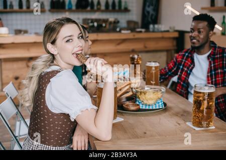 Fröhliche junge Frau, die Bratwurst isst, während sie mit bunten Freunden oktoberfest feiert Stockfoto