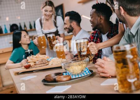 Selektive Konzentration der Kellnerin im traditionellen deutschen Kostüm, das Bier für multikulturelle Freunde im Pub serviert Stockfoto