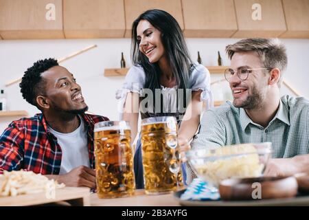 Fröhliche Kellnerin in traditioneller deutscher Tracht, die in Kneipe Bier für multikulturelle Freunde serviert Stockfoto