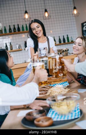 Attraktive Kellnerin in traditioneller deutscher Tracht, die ein Glas Bier hält, während sie in der Nähe von multikulturellen Freunden steht, die das oktoberfest im Pub feiern Stockfoto