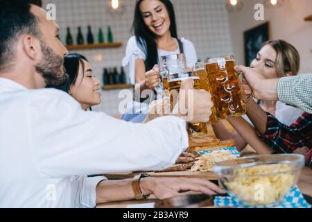 Selektive Konzentration der Kellnerin in der traditionellen deutschen Tracht, die Glas Bier hält, während sie in der Nähe von multikulturellen Freunden steht, die oktoberfest in p feiern Stockfoto