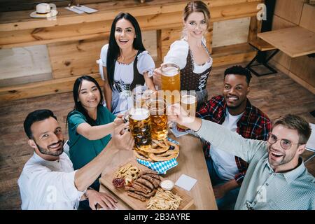 Attraktive Kellnerinnen in deutschen Trachten klatschen Bierbecher mit multikulturellen Freunden Stockfoto