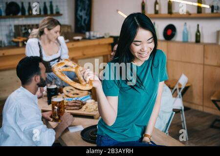 Selektiver Fokus des lächelnden asiatischen Mädchens, das Brezel hält, während er mit multikulturellen Freunden oktoberfest feiert Stockfoto