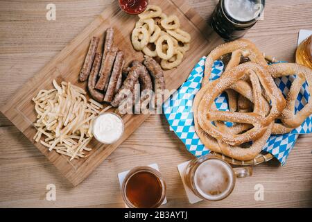 Blick auf Tassen mit hellem und dunklem Bier, gebratenen Würstchen, Zwiebelringen, pommes frites und Brezeln auf Holztisch im Pub Stockfoto