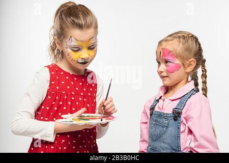 Niedliches Kind mit Katzen-Schnauze-Malerei auf dem Gesicht, mit Palette und Pinsel in der Nähe von Freunden mit bemalter Schmetterlingsmaske isoliert auf Weiß Stockfoto