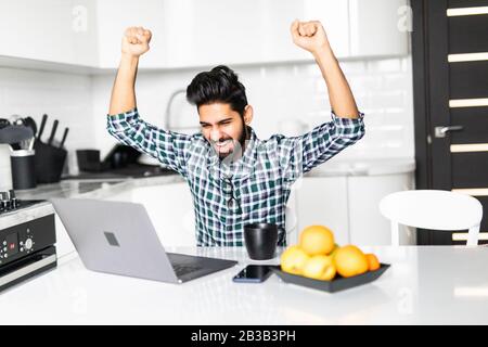 Junger indischer Mann feiert Erfolg bei der Arbeit an einem Laptop in der Küche Stockfoto