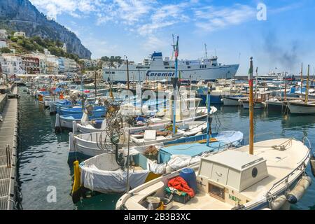 Insel CAPRI, ITALIEN - AUGUST 2019: Kleine Fischerboote im Hafen auf der Insel Capri. Im Hintergrund steht eine Autofähre. Stockfoto