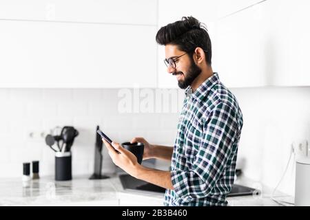 Der attraktive indische Mann liest Nachrichten auf dem Smartphone, das in der Küche steht Stockfoto