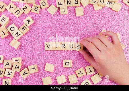 Ein Haufen Holzbuchstaben auf der Oberfläche eines rosafarbenen Hintergrundes und eine Hand bildeten das Wort Virus, selektiver Fokus. Stockfoto