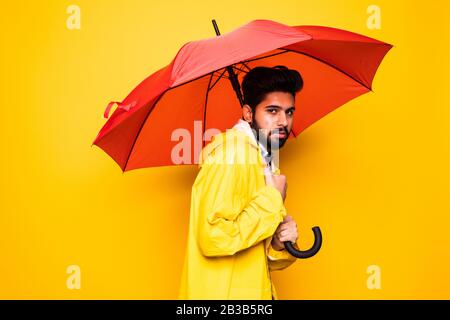 Junger, gutaussehender bärtiger indianermann in gelbem Regenmantel mit rotem Regenschirm aus Regen über orangefarbenem Hintergrund Stockfoto