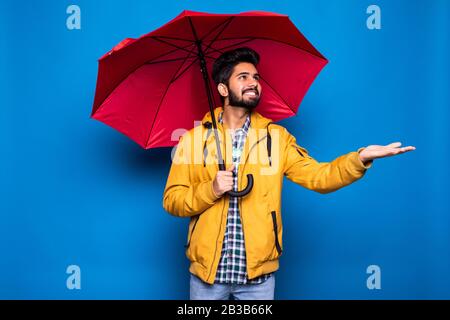 Junger, gutaussehender bärtiger indianermann in gelbem Regenmantel mit rotem Regenschirm aus Regen über blauem Grund Stockfoto