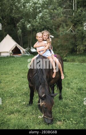 Reiten, zwei Kinder Mädchen, Schwestern, Reiten im Freien, lächeln und umarmen sich. Mädchen, die im Sommer auf einer Wiese reiten. Wigwam Stockfoto