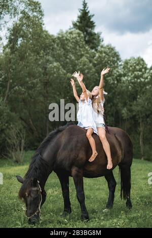 Zwei lustige Kinder Mädchen-Schwestern, die gemeinsam ein Pferd durch ein Feld reiten. Mädchen, die auf einem Pferd sitzen und Arme hochhalten, glückliches gehen im Freien Stockfoto