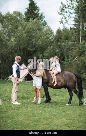 Fröhliche junge Familie mit zwei Kindern, die stylische Casual Boho-Kleidung und ein wunderschönes Pferd im Wald oder auf der Wiese trägt. Sommerzelt, indisch Stockfoto