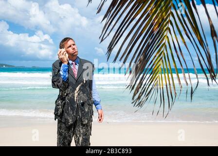 Gestrandeter Cadaway Geschäftsmann, der am Strand in seinem zerlumpten Anzug steht und ein imaginäres Gespräch auf seinem Shell-Telefon führt Stockfoto