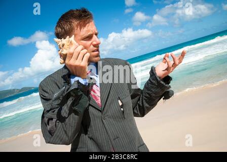 Gestrandeter Cadaway Geschäftsmann, der am Strand in seinem zerlumpten Anzug steht und ein imaginäres Gespräch auf seinem Shell-Telefon führt Stockfoto